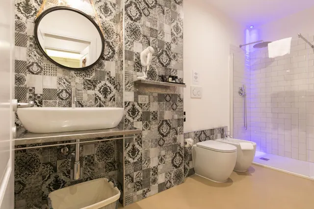 A bathroom with black and white patterned tiled walls, a vessel sink on a stone shelf, a toilet and bidet, and a walk-in shower with white tiles lit by blue light.