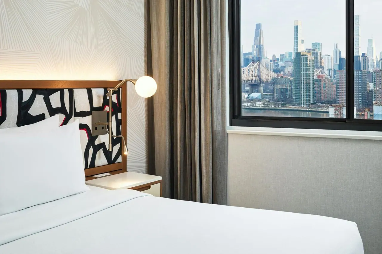 A made bed with white linens sits beside a window showing a city skyline and the Queensboro Bridge.