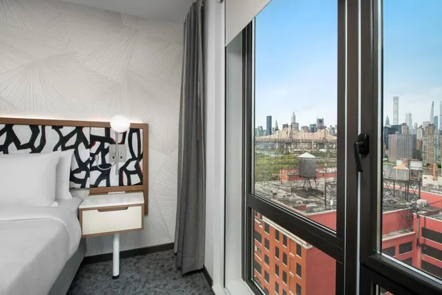 A bedroom with a made bed, a patterned headboard and a tall window looking out over a city skyline and a truss bridge.