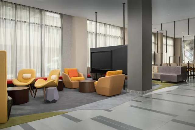 A lobby lounge with yellow armchairs, round wooden side tables, a wall-mounted television and a grey pillar beside large windows with sheer curtains.