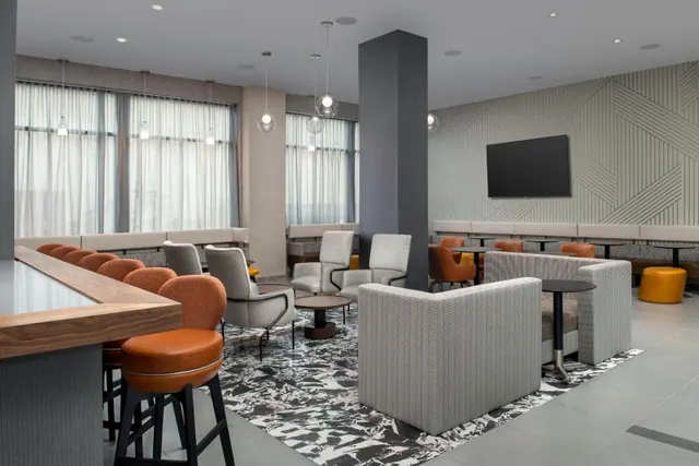 A hotel lounge with a wood-topped bar counter and orange stools, grey upholstered armchairs and sofas, booth seating along the wall, and a wall-mounted television.
