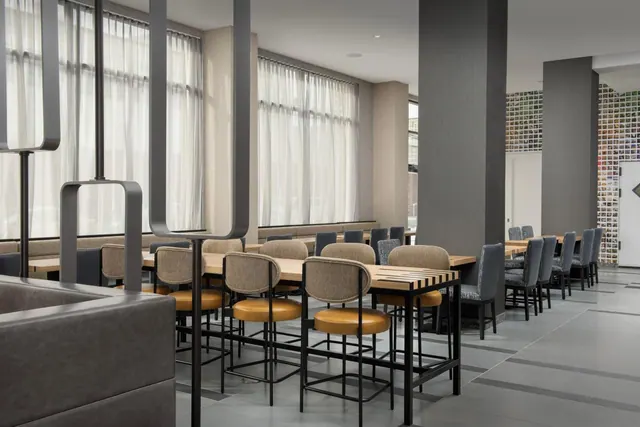 A dining area with high wooden tables, tan upholstered bar stools, tall windows with sheer curtains, and a brick wall at the back.