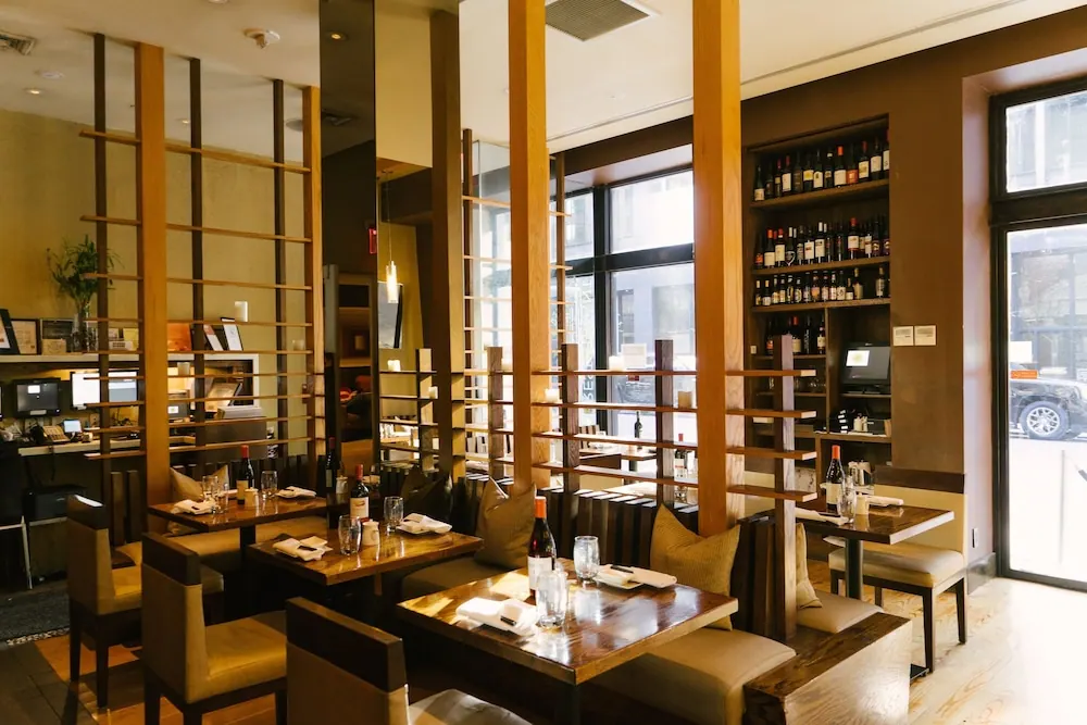 A restaurant dining room with wooden room dividers, tables set with glasses and wine bottles, wine shelves on the wall, and a window onto a street with a parked car.
