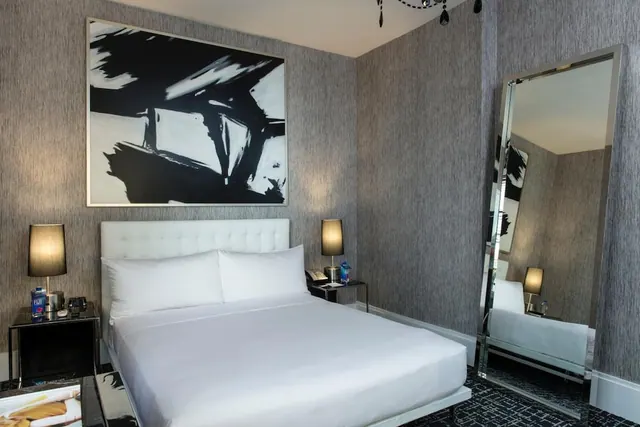 A bedroom with a made-up bed under a large black and white abstract painting, with a floor-length mirror and two bedside lamps.