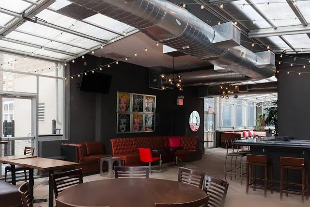 A bar lounge with red leather banquettes, a wall-mounted television, pop-art portrait prints, exposed ducts and a glass roof strung with lights.