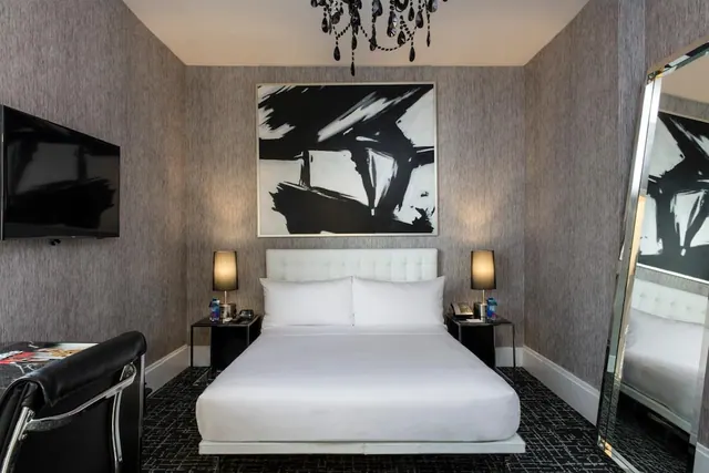 A bedroom with a made white bed, a wall-mounted television, a black leather desk chair, and a large black and white painting above the bed.