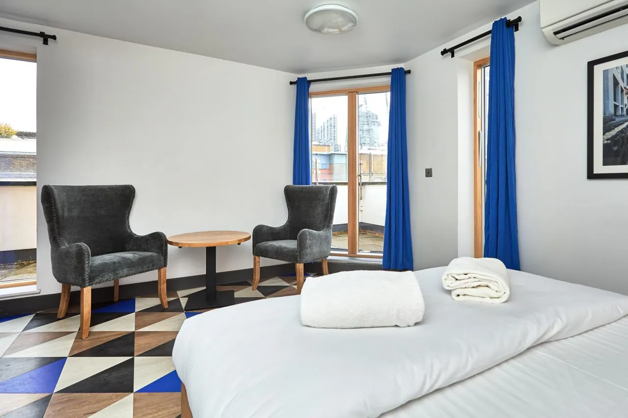 A bedroom with a made bed with white towels on top, two grey armchairs beside a small round wooden table, a geometric patterned floor, and blue curtains framing a glass door onto a balcony with buildings beyond.