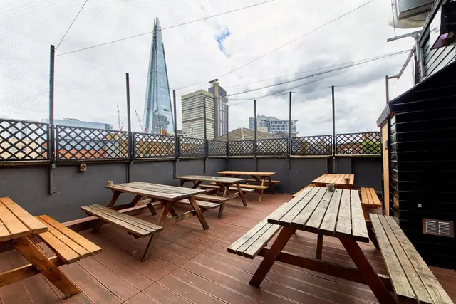 A rooftop terrace with wooden picnic tables and benches, with The Shard and other city buildings visible beyond the railing under an overcast sky.
