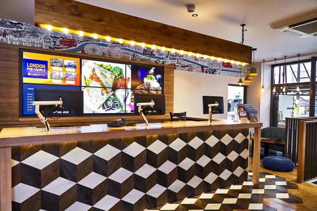 A hotel reception desk with a cube-patterned front sits below wall-mounted screens showing London pub crawls and food, with a London map wallpaper and bulb lights above.