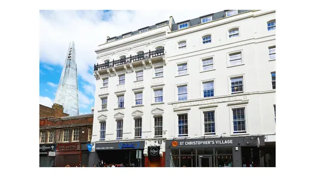 A white stucco building with a wrought iron balcony and street-level bars stands on a London street, with The Shard visible behind it.