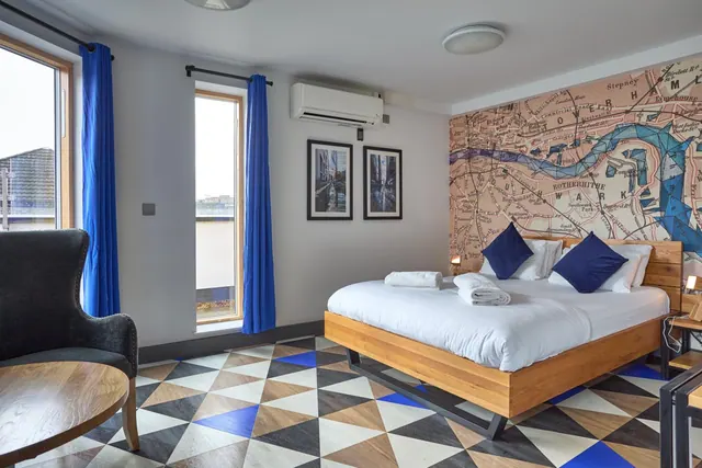 A bedroom with a double bed with blue cushions, a London map mural on the wall, blue curtains, a wall-mounted air conditioning unit and an armchair beside a round wooden table.