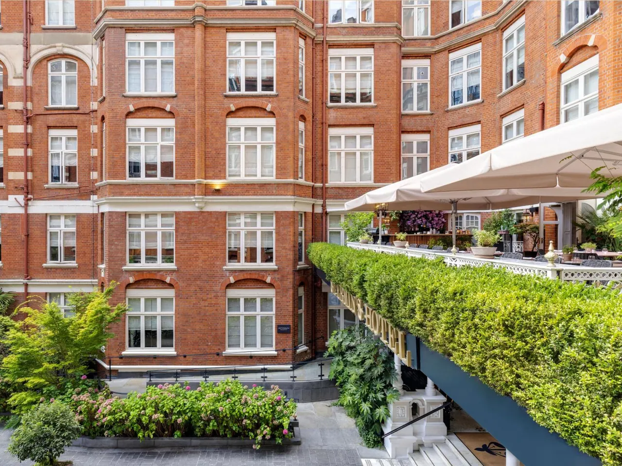 A red brick building facade with rows of windows rises above a courtyard with planters, a hedge-topped terrace and white parasols.