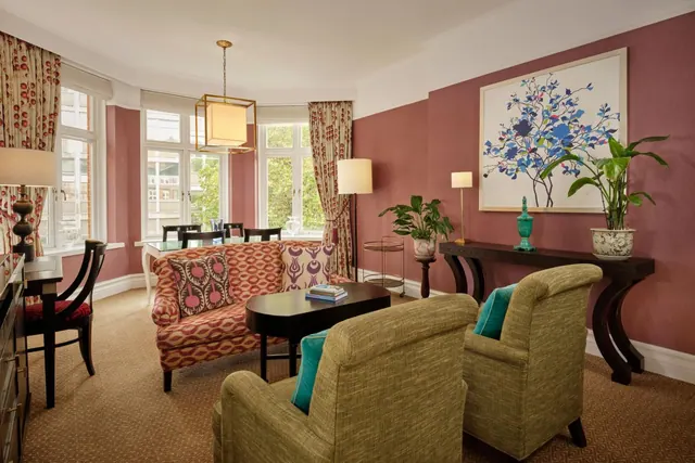 A living area in a hotel suite with a patterned sofa, two green armchairs, a coffee table, a dining table by a bay window and a large framed painting on a pink wall.