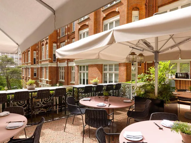 Outdoor terrace dining area with round pink tables, dark wicker chairs and cream parasols beside a red brick building.