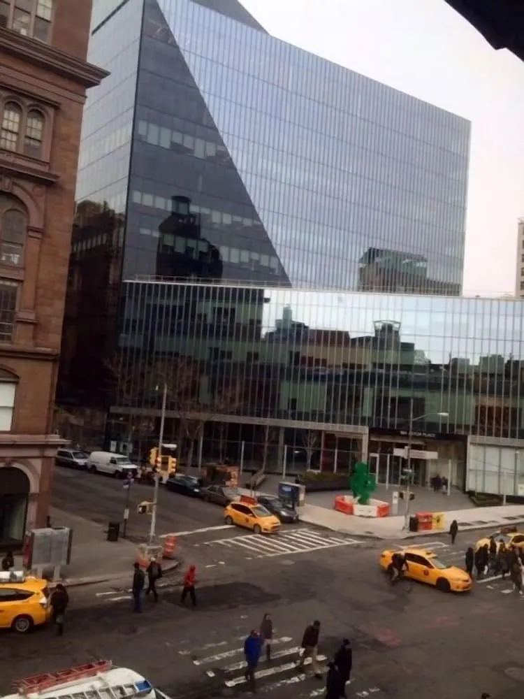 Photo taken from a window looking down on a city street with yellow taxis, pedestrians crossing at a crosswalk, and a large glass office building behind.