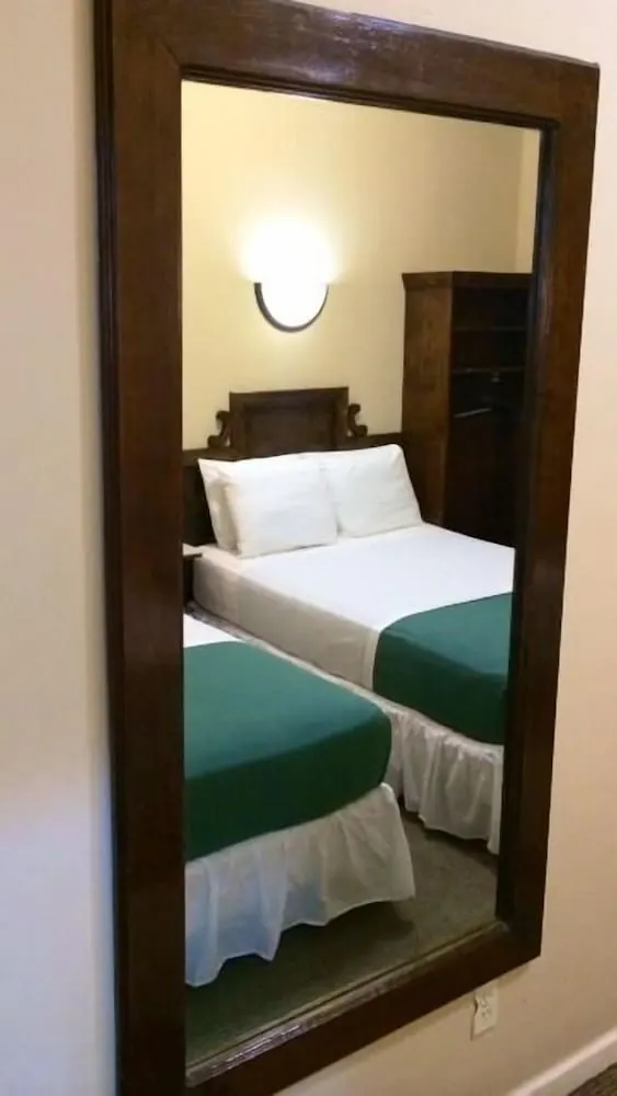 A wall mirror in a dark wooden frame reflects a hotel room with two beds with green bedspreads, a wall light and a wooden wardrobe.