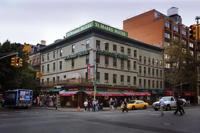 Street view of the St. Marks Hotel building on a corner, with a yellow taxi, a delivery van and pedestrians on the road.