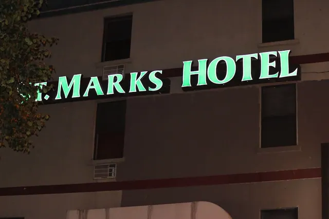 Night exterior of St. Marks Hotel showing its illuminated green sign above the upper windows, with a tree at the left edge and window air conditioning units below.