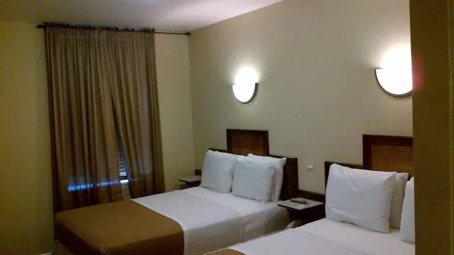 A bedroom with two beds with white bedding and dark wooden headboards, beside closed brown curtains and two wall lights.