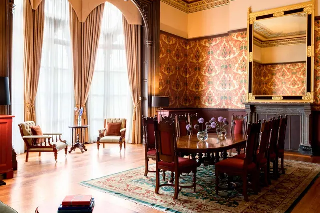 A formal room with a long wooden dining table and chairs, two armchairs by tall arched windows, patterned wallpaper and a gilt-framed mirror.