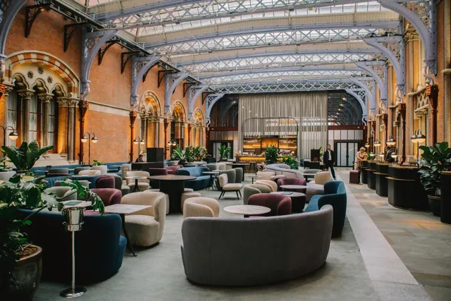 A large lounge with curved armchairs and small tables sits under an iron and glass roof, with brick walls, potted plants and a bar at the far end.