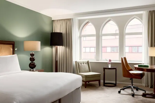 A hotel bedroom with a made bed, a green accent wall, an armchair, a desk and chair beside tall arched windows that look onto a brick building.