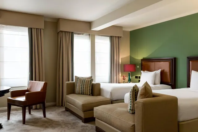 A hotel bedroom with two double beds with leather headboards against a green accent wall, an armchair and a upholstered seating area, and tall windows with sheer blinds.