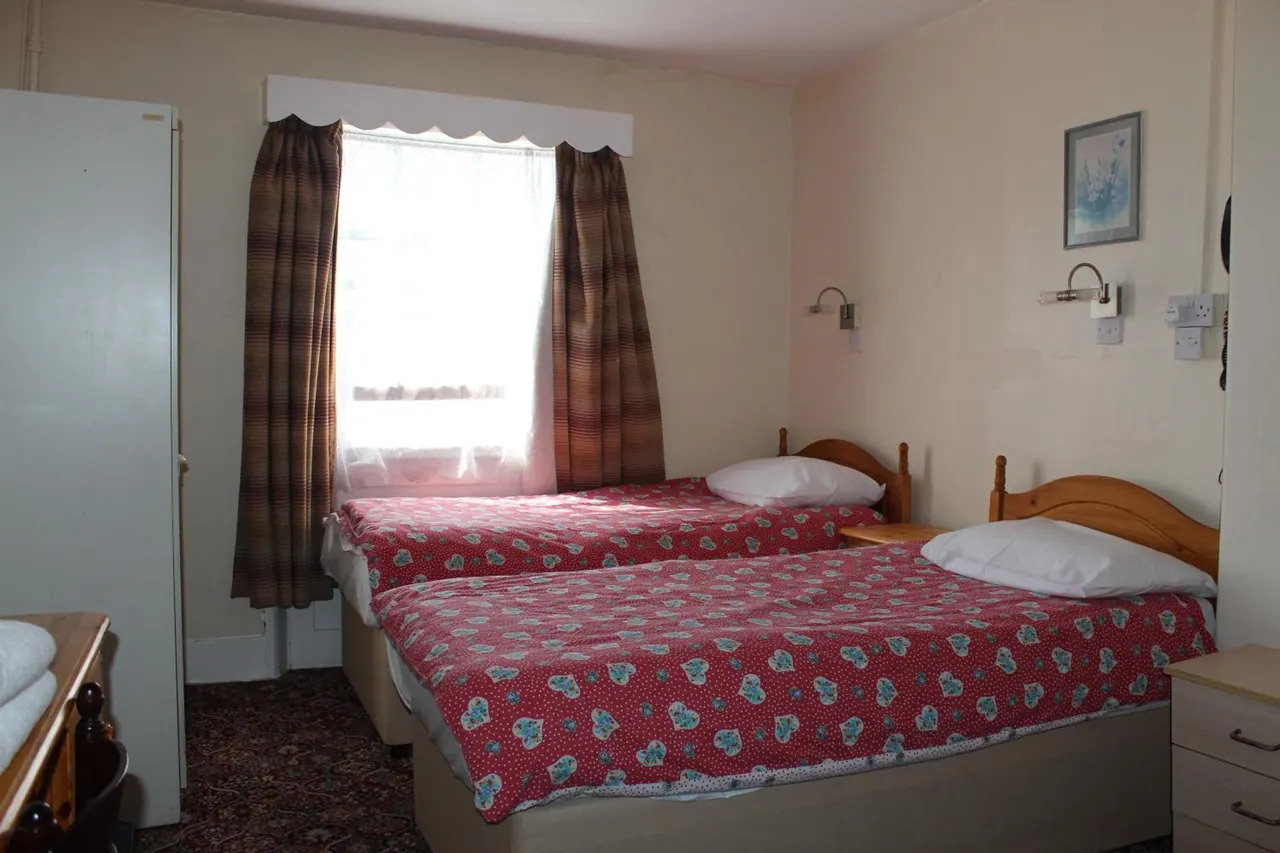 A bedroom with two twin beds with red floral bedspreads, a window with brown checked curtains, and a white wardrobe on the left.