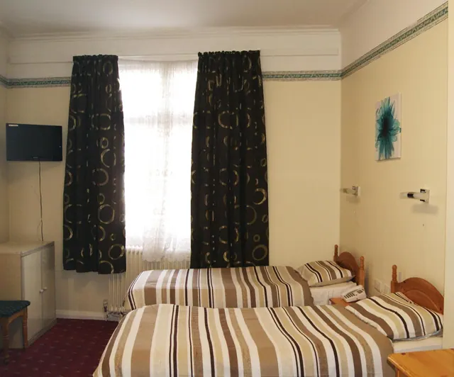 A twin-bedded hotel room with striped bedding, patterned dark curtains, a sheer-curtained window and a wall-mounted television.