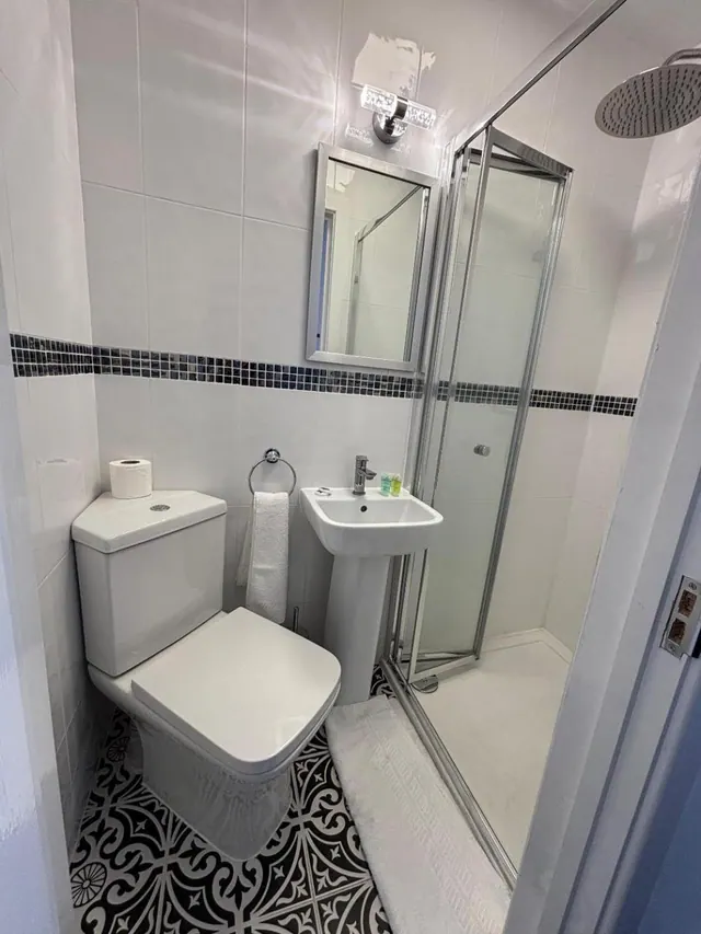 A small bathroom with a toilet, pedestal sink, wall mirror and a glass-enclosed shower with a rainfall head, on black and white patterned floor tiles.