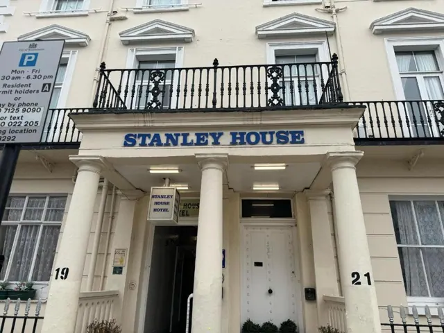 Front facade of Stanley House Hotel with white columns, a blue fascia sign, a white entrance door and an upper balcony with iron railings.