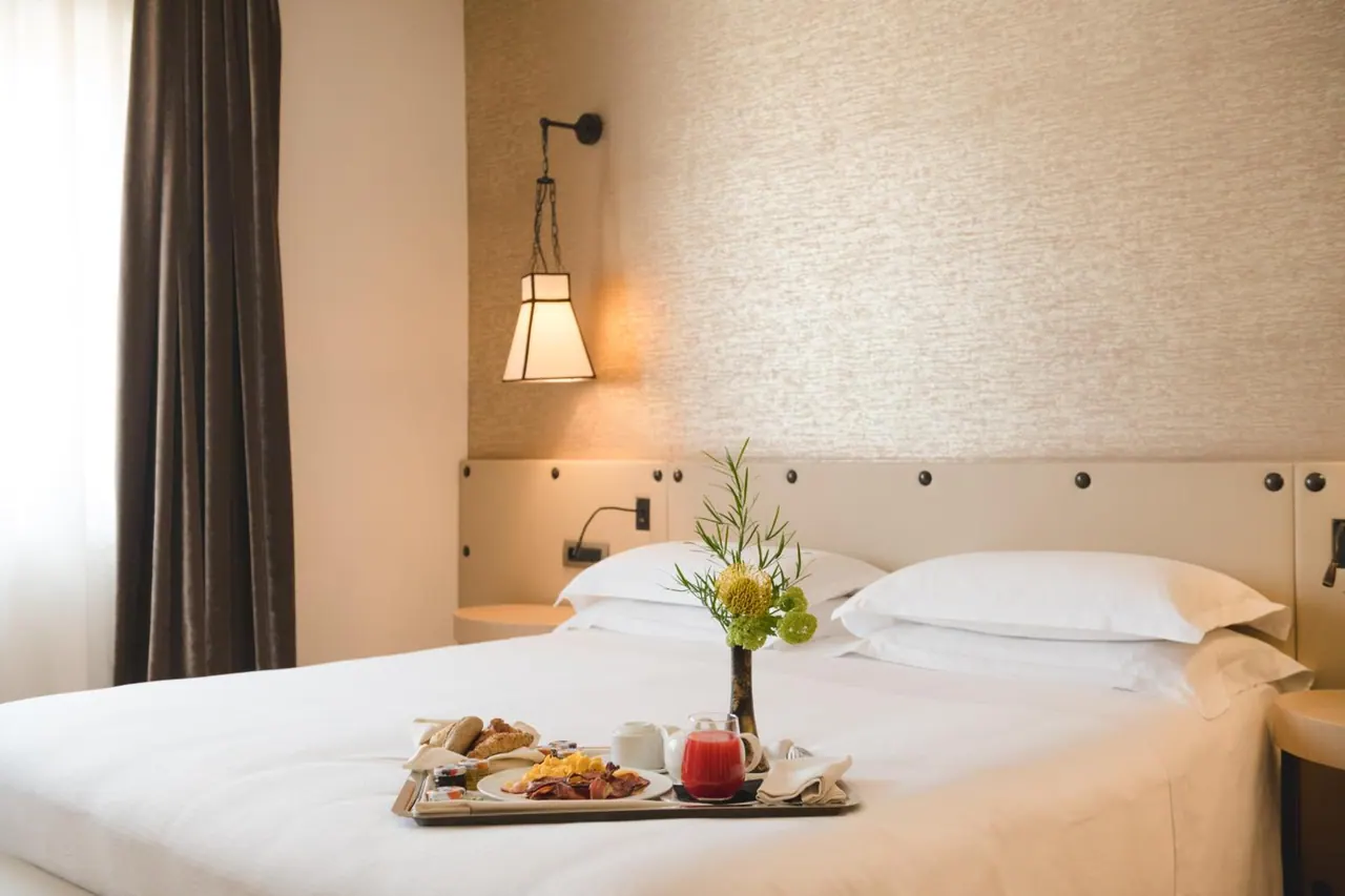 A double bed with white linen and pillows holds a breakfast tray with croissants, eggs, bacon, juice and coffee, with a wall lamp above the headboard.