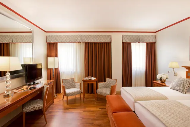 A hotel bedroom with two beds, a wooden desk with a television, two upholstered armchairs and a small side table by the windows.