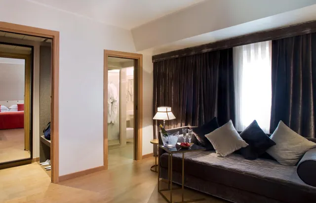 A living area in a hotel suite with a grey velvet sofa, a side table holding a champagne bucket, glasses and a fruit bowl, with an open doorway to a bathroom and a red bed reflected in a mirror on the left.