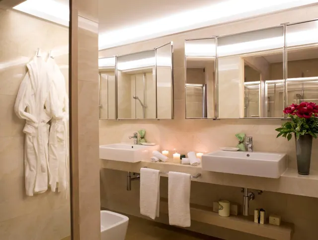 A bathroom with a double vanity under mirrored cabinets, white towels on a rail, lit candles, a bidet, and a glass-walled shower reflected in the mirrors.