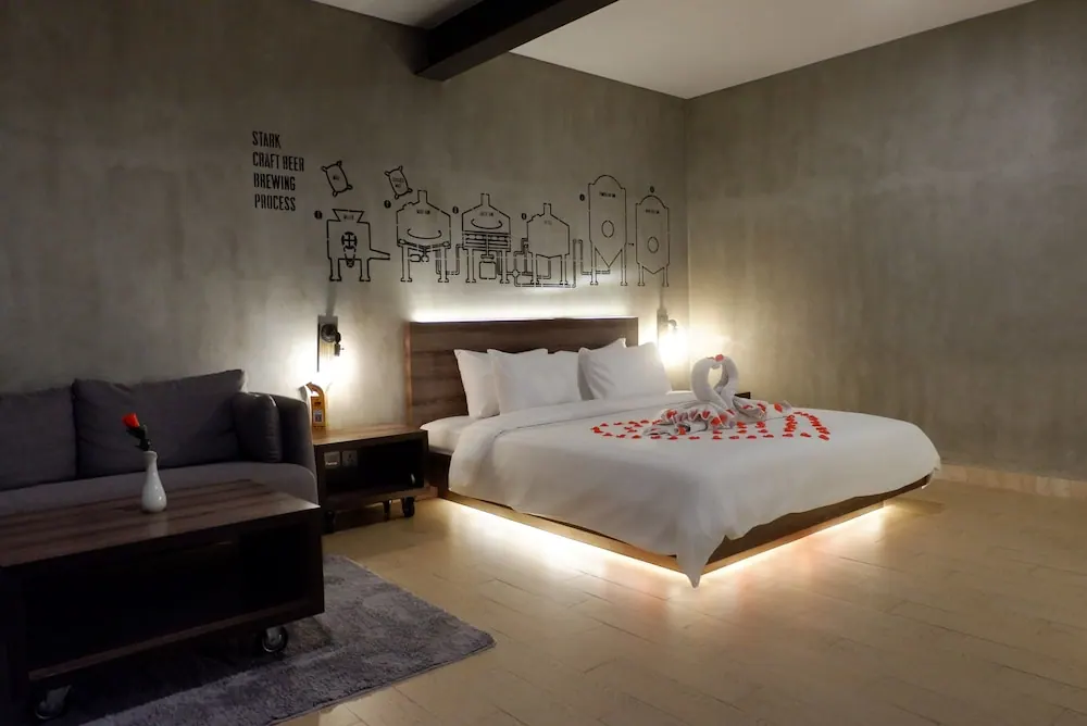 A bedroom with a king bed dressed with a towel swan and rose petals, a grey sofa and coffee table, and a wall drawing of a craft beer brewing process, lit by warm lights under the bed and headboard.