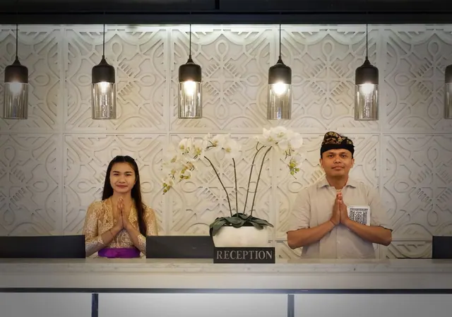 Two staff members in traditional Balinese dress stand behind a reception desk with a white orchid arrangement and a RECEPTION sign.