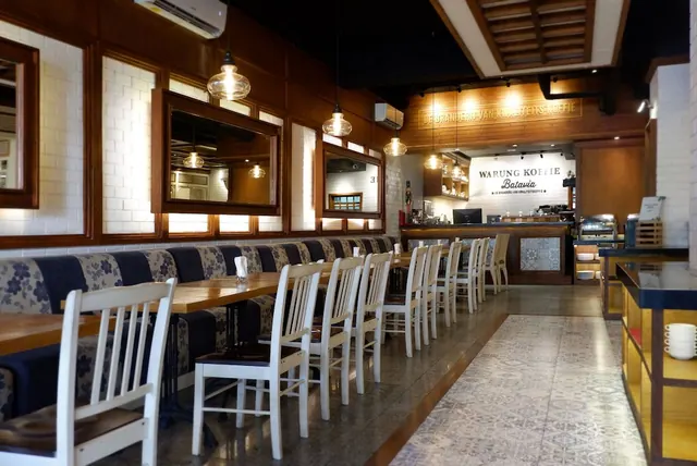 A dining room with wooden tables, white chairs and blue upholstered bench seating under pendant lights, with a counter and a Warung Kopie Batavia sign at the back.