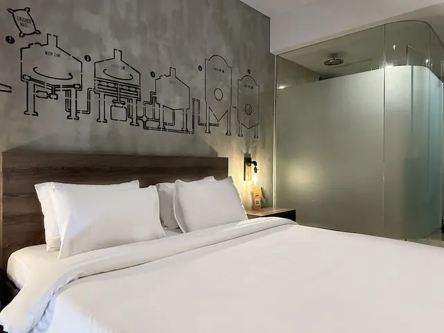 A bedroom with a made king bed with white linens, a wall decorated with a brewery equipment line drawing, and a frosted glass shower enclosure with a rain showerhead on the right.
