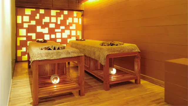 Two wooden massage beds with folded towels stand in a warmly lit treatment room beside a backlit wall of square panels.