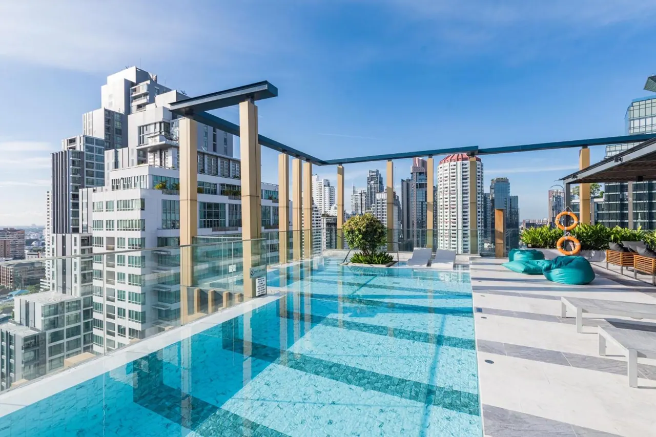 A rooftop infinity pool with a glass edge overlooks a city skyline, with sun loungers and teal bean bags on the deck.