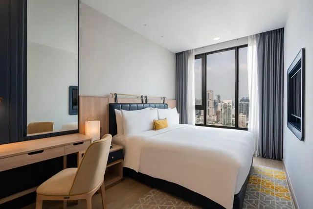 A bedroom with a made double bed, a wooden desk and chair, a wall-mounted television, and a window looking out over city high-rises.