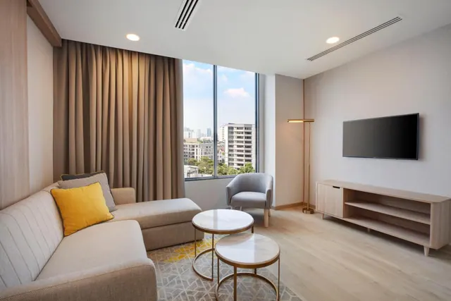 A living room with a grey sofa, yellow cushion, armchair, two round coffee tables and a wall-mounted television, with a window showing city buildings and trees outside.