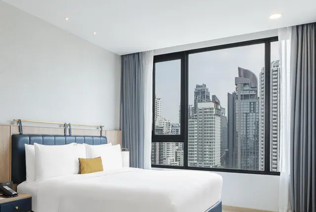 A made bed with white linens and a navy upholstered headboard sits beside a large window looking out onto high-rise buildings.