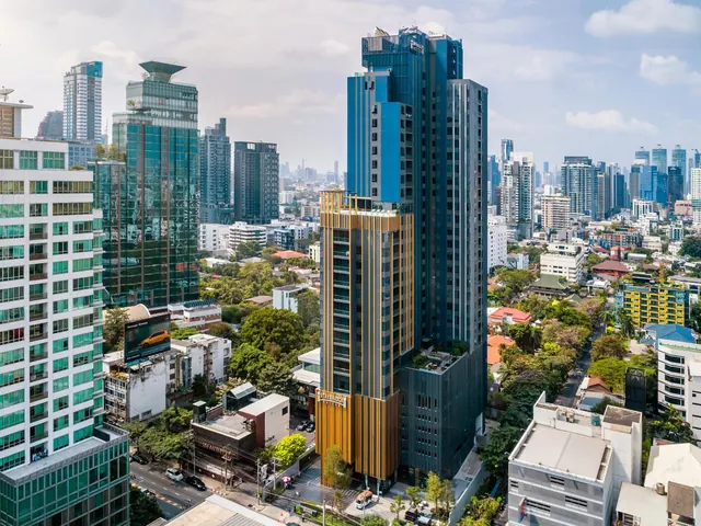 Aerial view of a tall blue glass tower with gold vertical fins, surrounded by other high-rises, low-rise buildings and trees in a city.