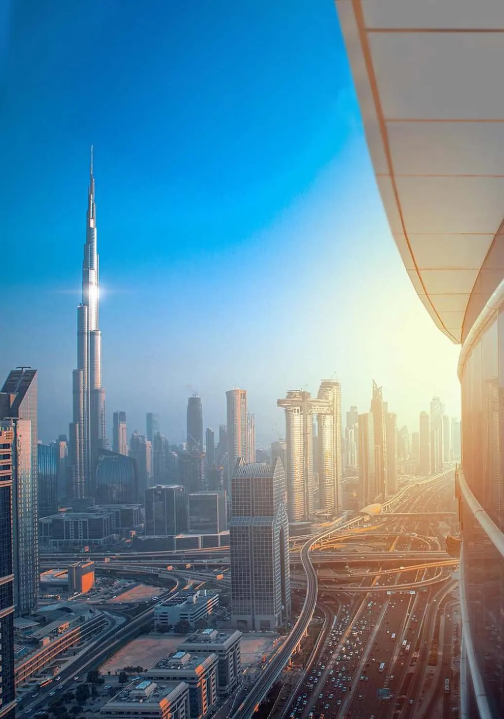 A view over the Dubai skyline toward the Burj Khalifa and a multi-lane highway interchange in warm low light, with a glass edge visible on the right.