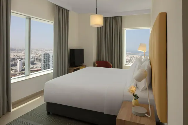 A bedroom with a made king bed, a television on a low cabinet, a red armchair, and large windows looking over a city skyline and water.