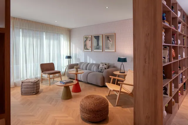 A living area with a grey sofa, wooden armchairs, round side tables, framed prints on the wall and a tall wooden bookshelf divider, with sheer curtains over a large window.