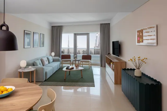 A living room with a light blue sofa, a round coffee table, a wall-mounted television and glass doors opening onto a balcony with a view of the Burj Khalifa and city towers.