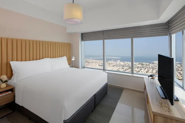 A king bed with white linen sits in a bedroom with large windows overlooking a city and the sea, with a wall-mounted television on a wooden console.